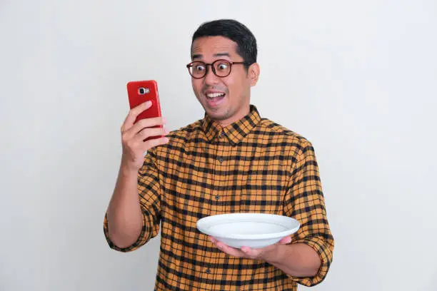 Adult Asian man looking to his handphone with amazed expression while holding dinner plate