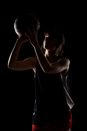 Female basketball player. Beautiful girl holding ball. Side lit half silhouette studio portrait against black background.