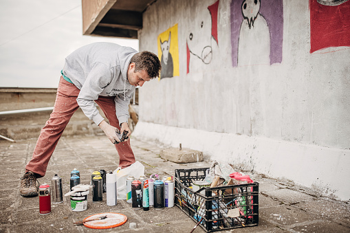 One male, male artist preparing spray paint for graffiti on the wall outdoors.