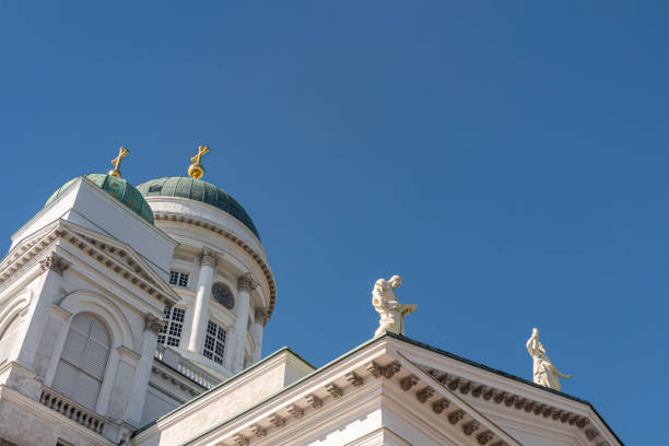 Helsinki, Finland - July 19, 2022: NE corner of white stone Cathedral against blue sky feauturing 2 green domes with golden pinnacles and 2 apostle statues: Philip carrying open book
