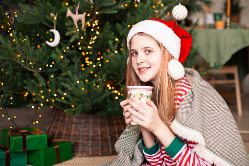 Girl with cup of drink wearing striped pajamas and Santa hat keeping warm under plaid near christmas tree. Happy teen waiting for Saint Nicholas. Selective focus.