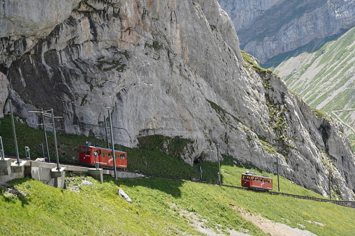 Pilatus, Switzerland 07 08 2022, Two red cogwheel trains between railway station Alpnachstad and mountain Pilatus Kulm. Behind are rocky cliffs of swiss alps. Track line are surrounded by green grass.