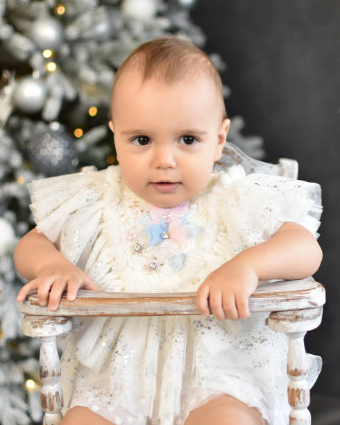 Baby 1 year old  sitting in chair with Christmas tree Baby 1 year old  sitting in chair with Christmas tree and lights on background in room. Merry Christmas and Happy New Year. Holiday season. baby new years eve new years day new year stock pictures, royalty-free photos & images