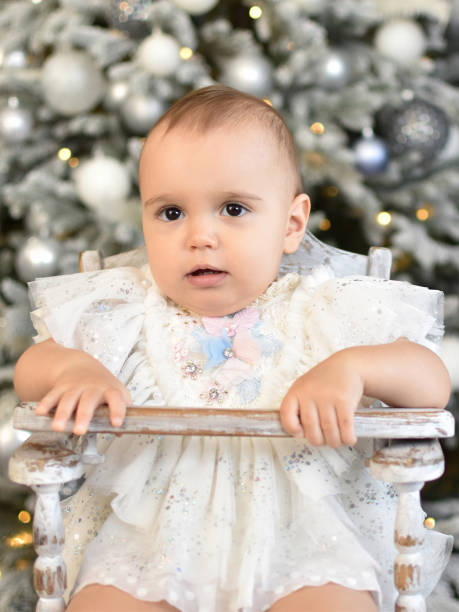 Baby 1 year old  sitting in chair with Christmas tree Baby 1 year old  sitting in chair with Christmas tree and lights on background in room. Merry Christmas and Happy New Year. Holiday season. baby new years eve new years day new year stock pictures, royalty-free photos & images