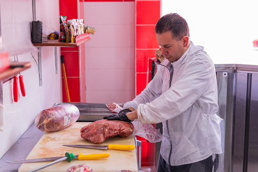 Latin male professional butcher of average age of 30 years old dressed in a white uniform is in the butcher shop for which he works cutting meat with portions
