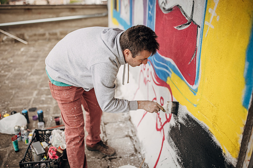 One male, male artist painting a graffiti with paintbrush on the wall outdoors.