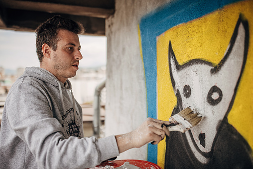 One male, male artist painting a graffiti with paintbrush on the wall on building top outdoors.