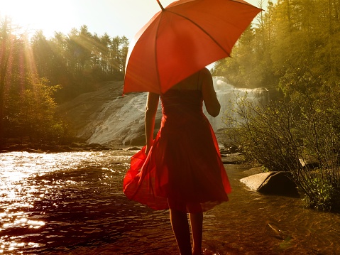 A young female with a red dress holding a red umbrella walking near a waterfall
