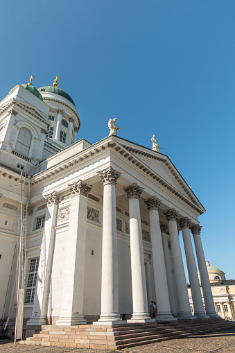 Helsinki, Finland - July 19, 2022: North side of white stone cathedral against blue sky. Columns support pediment with 2 apostle statues. 2 green domes with golden pinnacles. Escape ladder