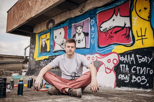 One male, male artist sitting by the graffiti that he painted on the wall on building.