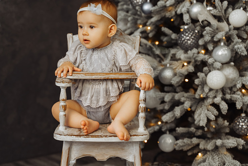 Adorable baby sitting in Baby high chair with Christmas tree and lights on background in room. Merry Christmas and Happy New Year.