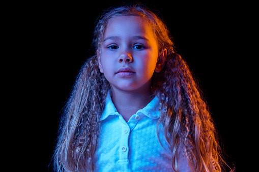 Resentment. Sad little girl, kid with long hair wearing white t-shirt expressing emotions isolated over black background in neon light. Concept of children emotions, fashion, beauty, back to school, ad