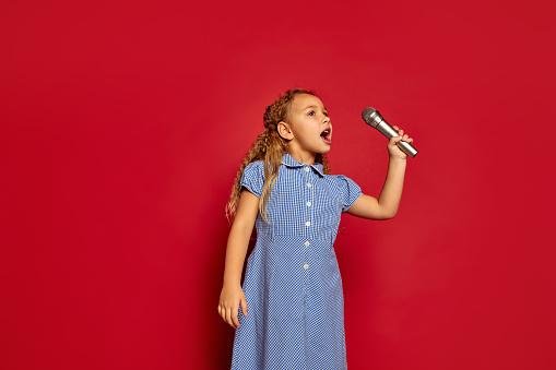Cute little kid girl isolated on red studio background. Human emotions, facial expression. Concept of childhood, children, lifestyle. Mock up copy space for ad. She listen music, sing song in microphone