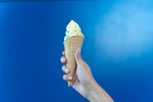 woman hand holding a cone of yellow soft serve ice cream on blue background.