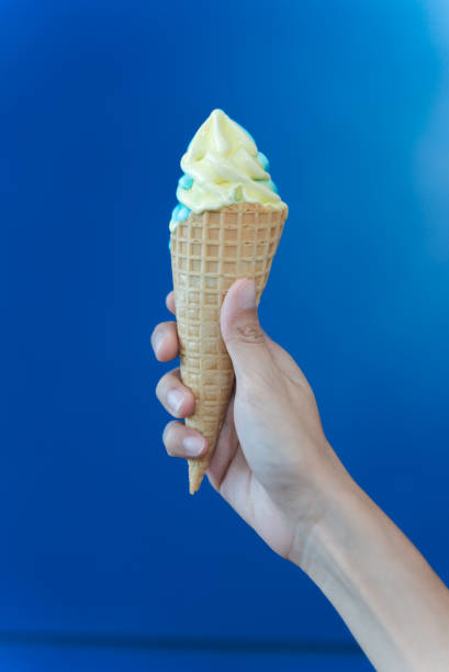 istock Female hand holding yellow ice cream cone on blue background. 1438691927