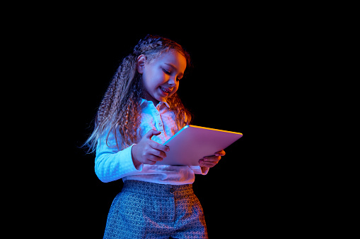 Pretty nice-looking little girl with laptop isolated over dark background in neon light. Concept of human emotions, beauty, kids fashion, children, school and ad. Copy space. Sales, black Friday