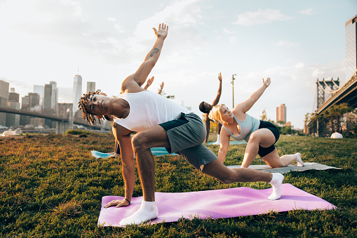 Group of young people attending to a yoga class outdoors at sunset with New York cityscape on their background. They are meditating and relaxing.