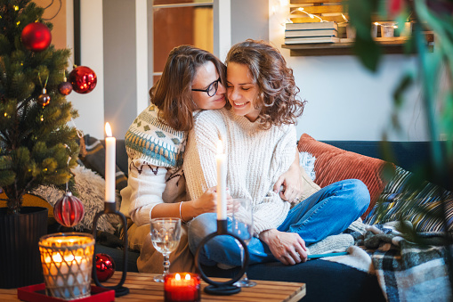 Young adult happy lesbian couple sitting on couch at home hugging and celebrating christmas and new year eve