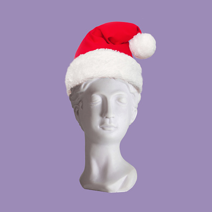 Female antique statue's bust in Santa hat on her head isolated on purple color background. Trendy collage in magazine surreal style. 3d contemporary art. Modern design. Christmas and New Year concept