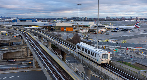 New York City, USA - April 8, 2022: JFK International Airport, one of the busiest in the world, is active even in stormy weather in early morning. Airtrain is going to the terminal. The British Airways with Alaska Airlines airplane is parked to a jet bridge in Terminal 7 for boarding, servicing, and baggage loading.