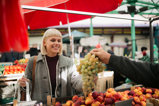 Happy young woman with blond hair visiting a local farmer's market, eating healthy and buying some grapes from a local vendor