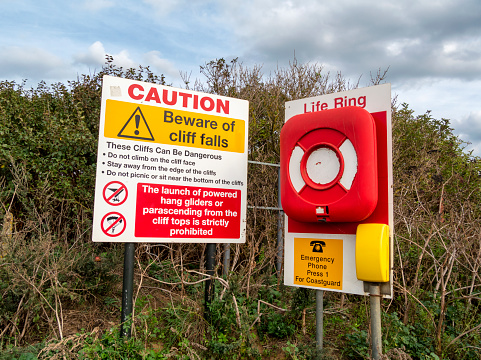 A notice warning of cliff falls and the dangers of climbing on the cliffs beside a life ring and Coastguard telephone at Old Hunstanton in Norfolk, Eastern England.