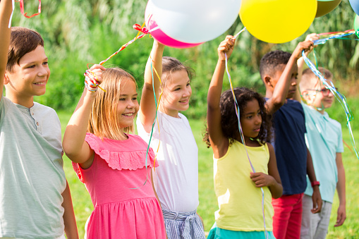 Portrait of cheerful emotional preteen boys and girls of different nationalities with colorful toy balloons in hands posing on green lawn in city park on sunny summer day