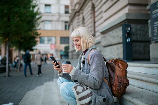 Side view of a happy young adult female university student sitting in front of the university building, having a cup of coffee and using a smart phone, enjoying herself