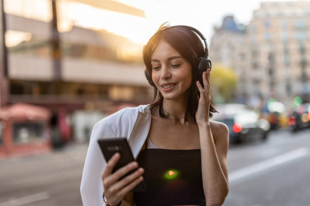 a young hispanic woman walks down a city street listening to music on wireless headphones and using a mobile phone - city life audio imagens e fotografias de stock