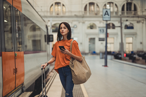 A young beautiful Spanish woman is using a mobile phone at the train station while waiting to enter the train car, she is dressed in an orange T-shirt, a casual wardrobe, she is going on a tourist trip