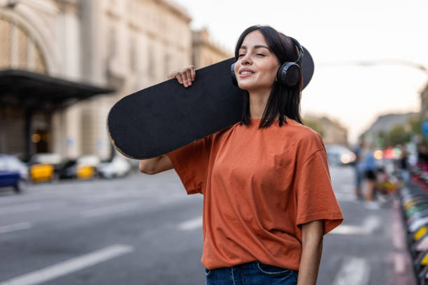 une jeune femme espagnole avec une planche à roulettes marche dans la rue - headphones relaxation outdoors caucasian photos et images de collection