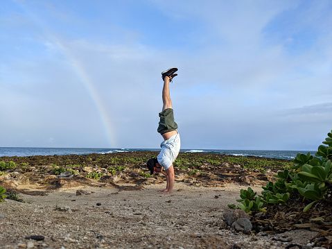 Man does Handstand on coral rocks on the beach with rainbow in the sky as wave crash in the distance on the North Shore of Oahu, Hawaii.