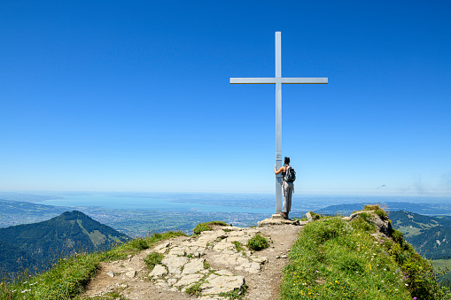 Young woman on top of a mountain (Moerzelspitze) holding the summit cross looking at Lake of Constance in the background