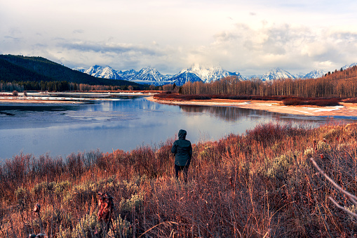 A hiker stands in a field on the side of a hill enjoying the beautiful views of the Grand Tetons reflecting in Lake Jackson.