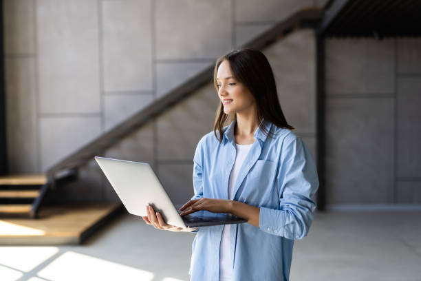 istock Attractive smiling young brunette woman using laptop computer while standing in a stylish living room, 1438749645