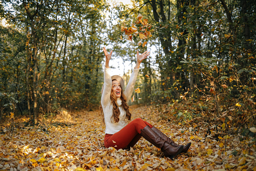 Positive curly haired woman throwing dry leaves in picturesque autumn forest with colorful trees on sunny day