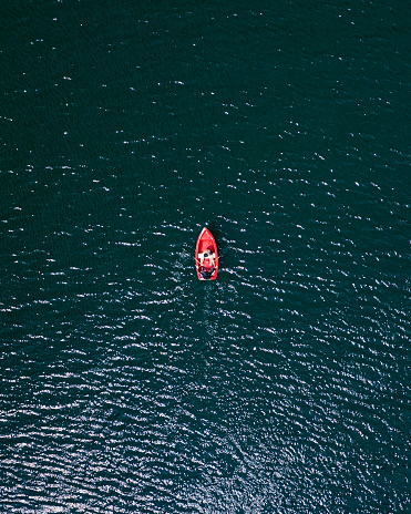 A red boat in the middle of a blue lake steered by two people. A beautiful red kayak on an open lake, open water, open sea. Two people steer the boat on calm water. Minimalistic photo.