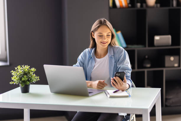 istock Young woman working from home using smart phone and computer, woman's hands using smart phone in interior, at home workplace using technology. 1438753454