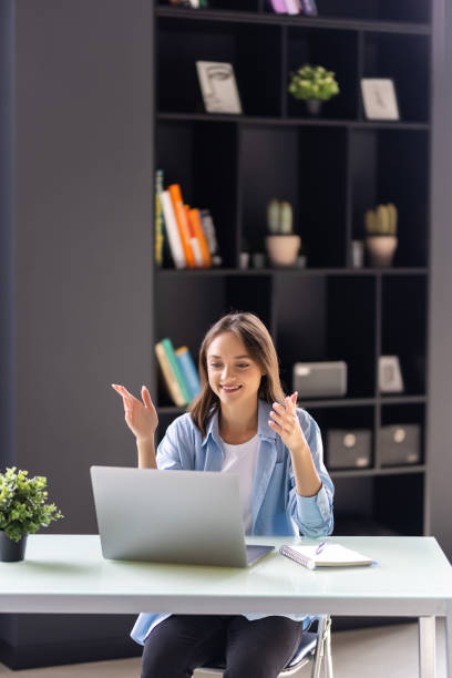 istock Young business woman making video call with computer while talking with earphone sitting in modern startup office. 1438753906