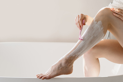 Cropped view of woman depilating shins over ceramic bathtub. Female shaved leg with pink plastic razor. Lady using epilation gel foam for sensitive skin. Pampering, moisturizing, self care in bathroom