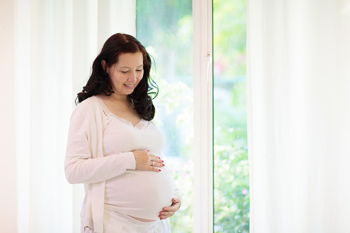 Pregnant woman at home. Asian expecting mother in white bedroom. Healthy pregnancy. Young female looking at baby sonogram preparing for birth. Maternity and prenatal care.