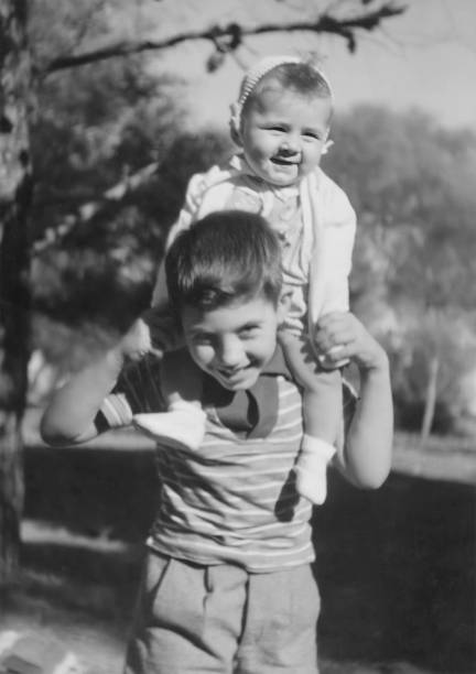 black and white image taken in the 50s: little boy carrying his little sister on his shoulders - family child portrait little girls imagens e fotografias de stock