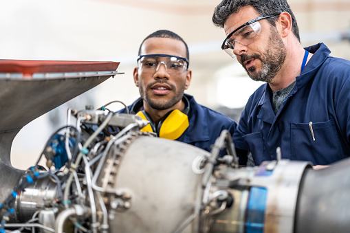 Two aircraft engineers in protective eyewear discussing jet turbine repairing, close view. Experienced mid adult male aviation mechanic teaching coworker examining helicopter