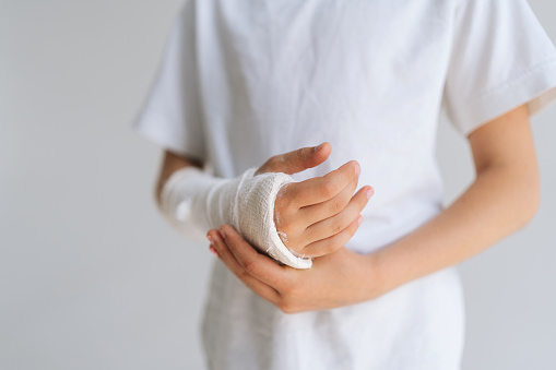 Close-up cropped shot of unrecognizable little girl with broken hand wrapped in white plaster bandage touching injured forearm on light isolated background. Concept of child insurance and healthcare.