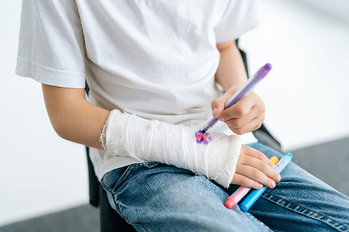 Close-up cropped shot of unrecognizable little girl drawing cute image on cast of broken arm wrapped in white plaster bandage on white isolated background. Concept of child insurance and healthcare.