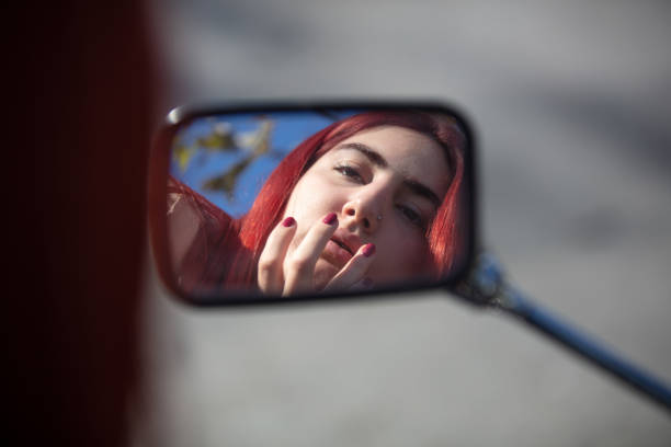 Young girl looking at her makeup in the mirror.\nThe girl uses a motorcycle mirror to look at her makeup on the street.