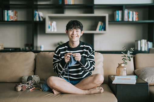 Asian Chinese LGBTQIA gay man looking at camera knitting at home sitting on sofa in living room