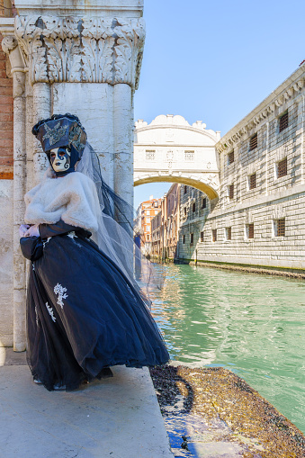 Venice, Italy - March 01, 2022: Woman dressed in traditional costume stand in front of the Bridge of Sighs, part of the Venice Mask Carnival, Veneto, Italy