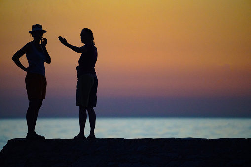 Silhouettes, side, back view of two women standing on the rock, discussing while looking at sunset on Adriatic coast in Croatia.