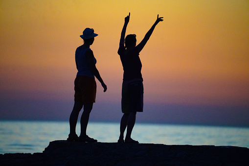 Silhouettes, side, back view of two women standing on the rock and looking at sunset on Adriatic coast in Croatia.